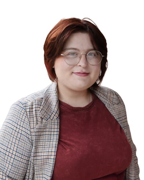 Close-up of a young woman with short red hair, round glasses, a nose ring, wearing a burgundy shirt and a plaid blazer, smiling.
