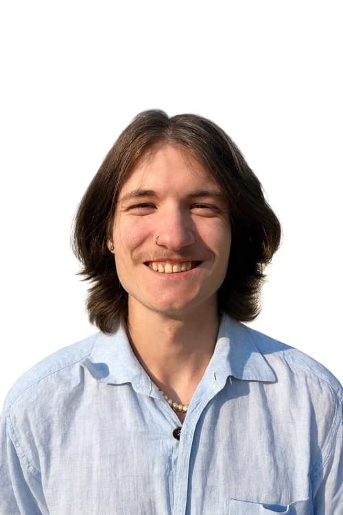 Portrait of a smiling young person with shoulder-length brown hair, wearing a light blue button-up shirt, a pearl necklace, and a nose ring, against a plain white background.
